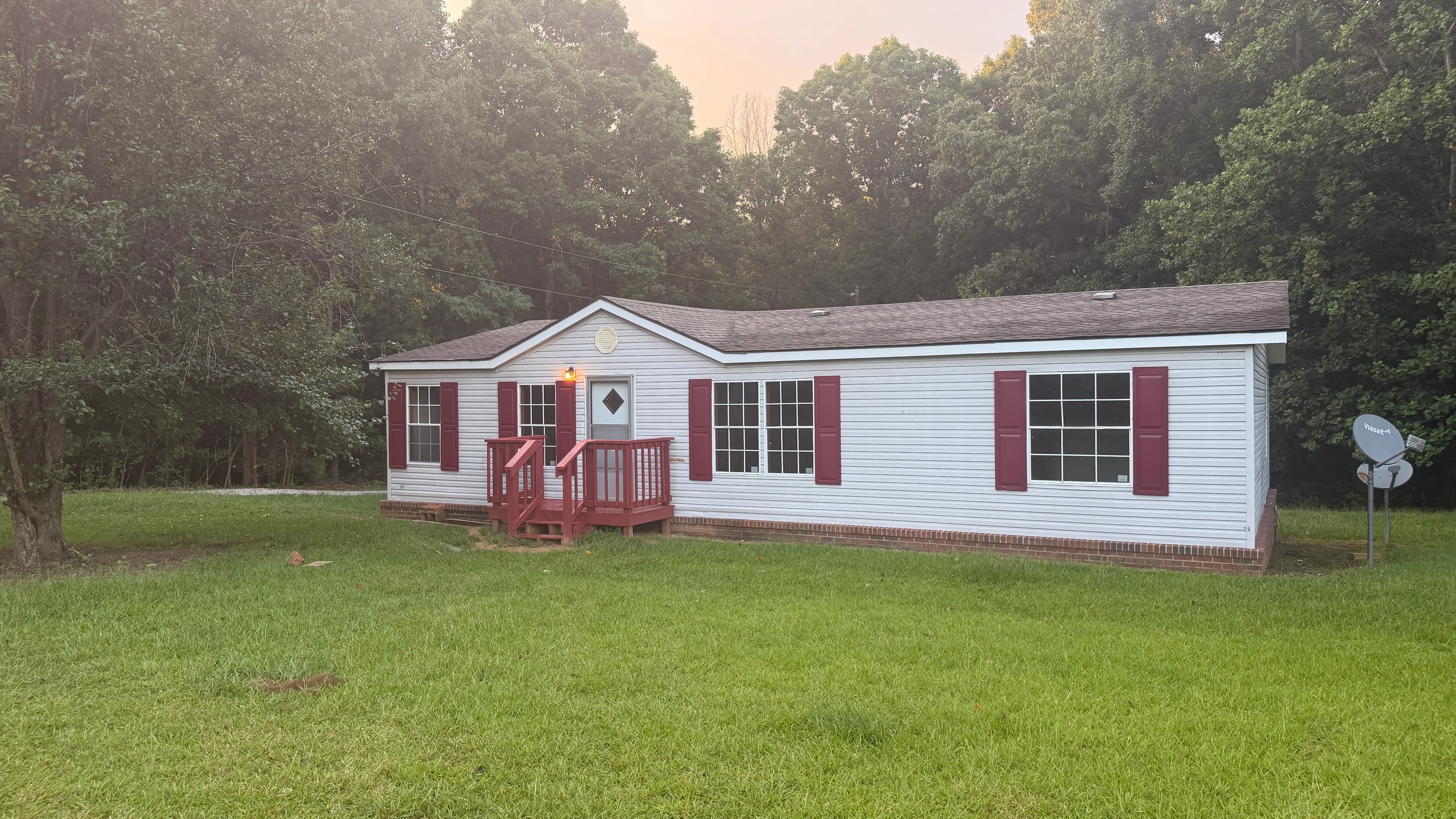 White vinyl siding mobile home with red shutters, brick foundation, and red wood entry deck