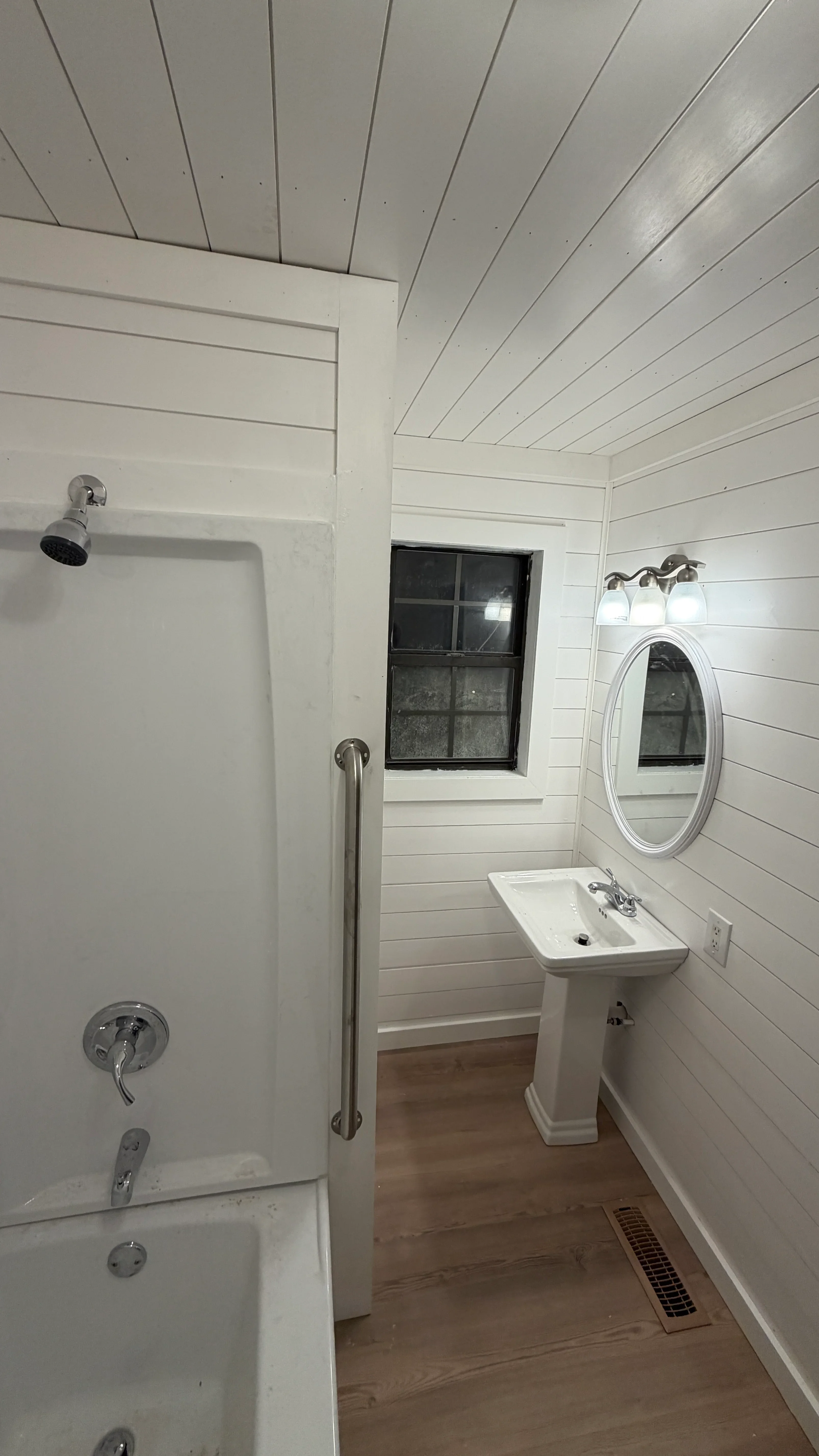 White shiplap bathroom with pedestal sink, shower, oval mirror, and vaulted ceiling